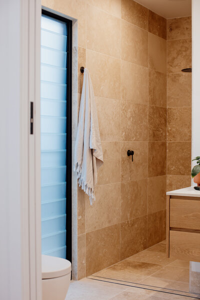 Minimalist bathroom with beige tiles, a towel hanging near the shower, and natural light streaming through a frosted window—an elegant creation by a Newcastle Kitchen and Bathroom Designer.