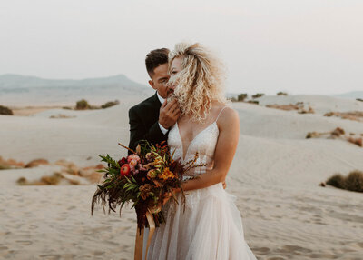 Bride with curly hair holding boho wedding bouquet while hugging the groom at the Sand Dunes taken by Phoenix wedding photographer, Kaylie Miller.