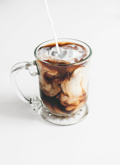 Glass mug filled with iced coffee as cream is poured in, creating a swirling effect, symbolizing a refreshing and energizing moment of creativity and focus