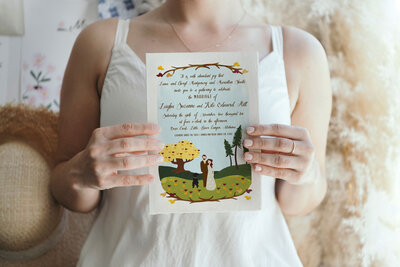 Whimsical wedding invitation with hand-drawn illustration of the bride and groom, fur-baby, and woodsy fall details.