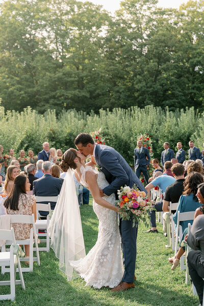 Bride and groom sharing their ceremony kiss at an outdoor Michigan orchard wedding with guests seated behind them.
