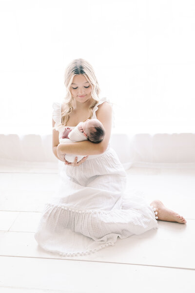 A young mother is sitting on the floor while holding her newborn baby and looking down at him.