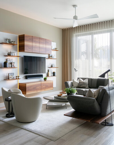 Living room decorated with neutral colours. The photo looks into the living room on an angle, with the wooden floating shelf tv unit as the focal point. The room has white fabric chairs, and a dark grey couch.