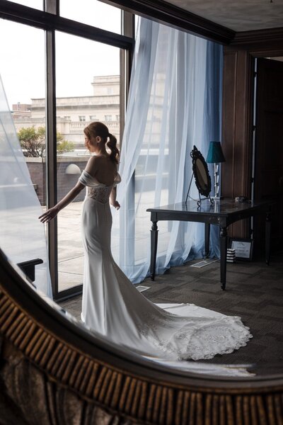 Bride standing gracefully by a floor-to-ceiling window at JPS Events, her fitted gown with lace details trailing behind her as soft light filters through sheer blue curtains.