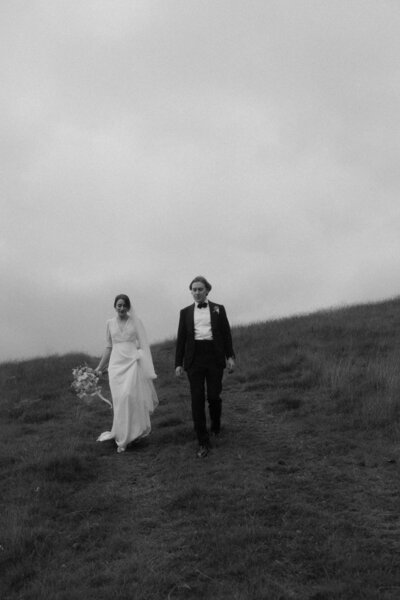 Black and white photo of a bride and groom walking across a grassy hillside, evoking timeless editorial (upper house estate derbyshire) elegance in UK countryside.