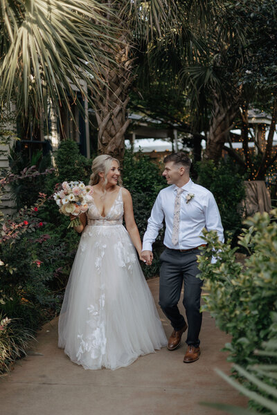 A newlywed husband and wife hold hands while walking through the gardens at the River Road and Jasmine House in Columbia, South Carolina. They're smiling at each other.