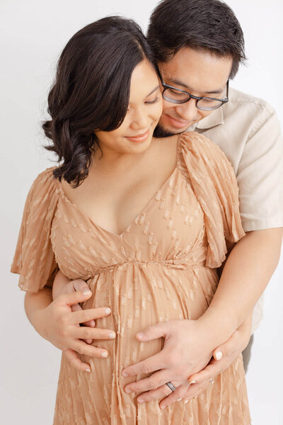 Pregnant Mom in beautiful taupe dress. She is looking down and holding her belly. Dad is standing behind her with his head on her shoulder and he is hugging her belly and also looking down.