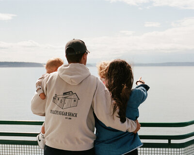 A family of four, seen from behind, embraces while looking at a vast, calm ocean. Both children are held by an adult looking towards the horizon.