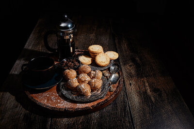 A cozy and inviting image of cookies, pastries, and a French press coffee set on a rustic wooden table. Photographed by Mica McCook, this scene evokes warmth and comfort, perfect for a relaxing coffee break.