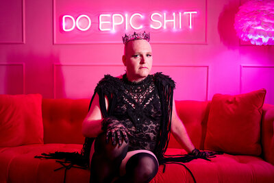 A person in lingerie sitting on a couch with a neon sign behind them that reads "Do epic shit".