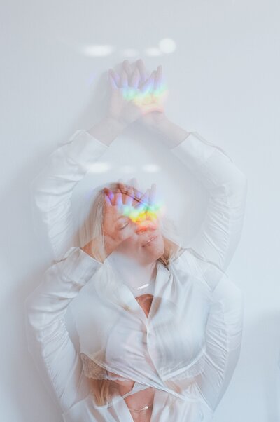Double exposure of woman with blonde, wavy hair and silk, white top with rainbow and disco ball reflections on her face stretching her arms above her.