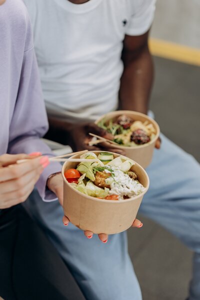 Two people holding bowls of food in takeout containers