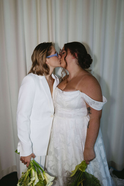 LGBT wedding couple sharing a kiss in elegant white attire, framed by beautiful curtains and holding bridal bouquets