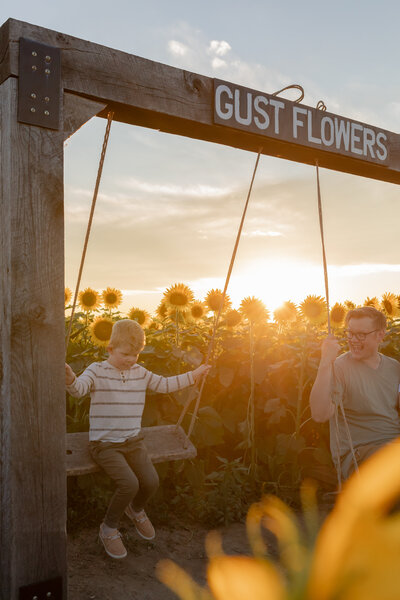 Boy swings on a wooden swing with father in the middle of a sunflower field at Gust Flower and Produce Farm in Ottawa Lake Michigan
