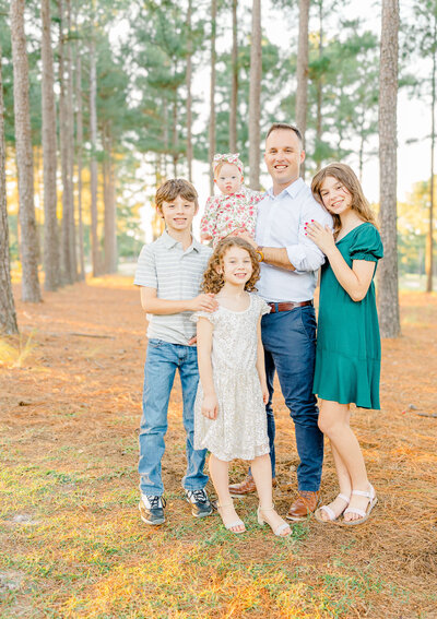 A Fayetteville NC family photographer captures a heartwarming moment as a family of five poses among the tall, majestic trees. The group, casually dressed, includes a man cradling a baby, and three children. Sunlight dances through the branches, highlighting the pine needle-covered ground.
