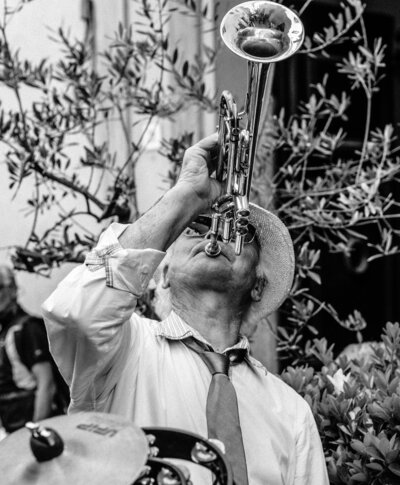 Schwarz-weiß-Foto eines älteren Straßenmusikers in Italien, der leidenschaftlich Trompete spielt – mit Strohhut, Hemd und Krawatte, im Hintergrund Blätterzweige und im Vordergrund teilweise sichtbare Trommel