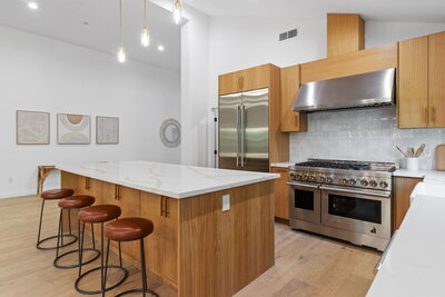 Bright modern kitchen featuring natural wood cabinetry, waterfall-edge island with white quartz countertops, leather bar stools, and stainless steel appliances