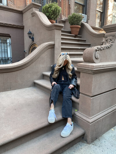 A person with long blonde hair sits on elegant brownstone steps, wearing a black outfit and white sneakers. They look down, creating a relaxed and contemplative atmosphere.