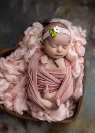 newborn baby wrapped in pink fabric wearing a pink floral headband asleep in a heart shaped wooden bowl