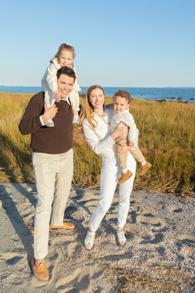 A dad, mom, and their two young children smile at the camera as Golden Hour sunlight lengthens their shadows behind them. In the background, the viewer can see the beauty of the grassy Connecticut coastline, and glistening blue water.