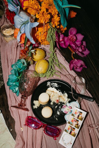 A close up of a wedding reception table with a plate of cookies and cake, photo strip, colorful florals, and glitter covered lemons