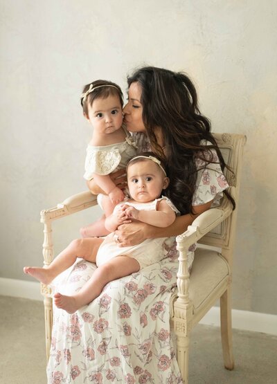 Mother in floral dress holds twin baby girls, kissing one while sitting on a vintage-style chair.