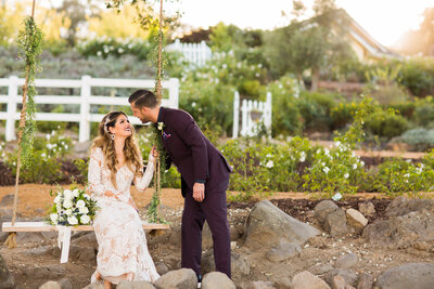 Bride sits on a swing bench and smiles at her Groom as he leans towards her for a kiss