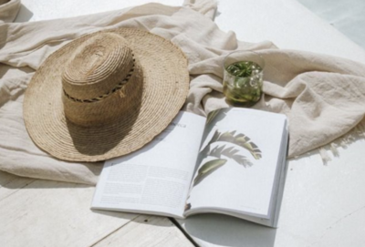 Flat lay of a straw sunhat, open botanical book, green drink, and soft neutral blanket by a bright white poolside surface