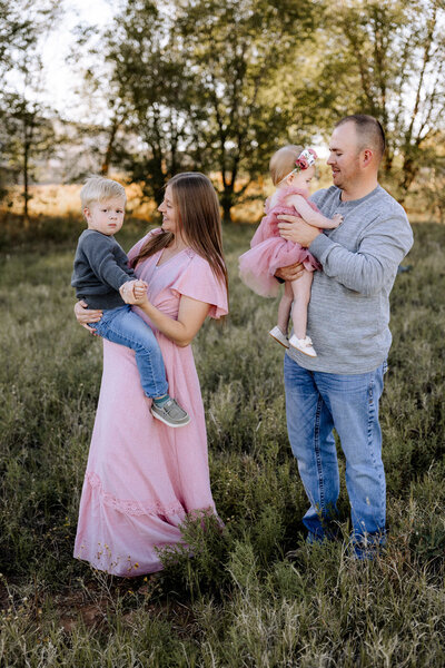 A joyful family portrait taken outdoors at sunset, featuring a mother in a black dress holding her young son, a father in a cowboy hat playfully holding his son upside down, and their other son standing between them, all dressed in coordinating black tops and denim. Captured by Mary Wiebe Photography