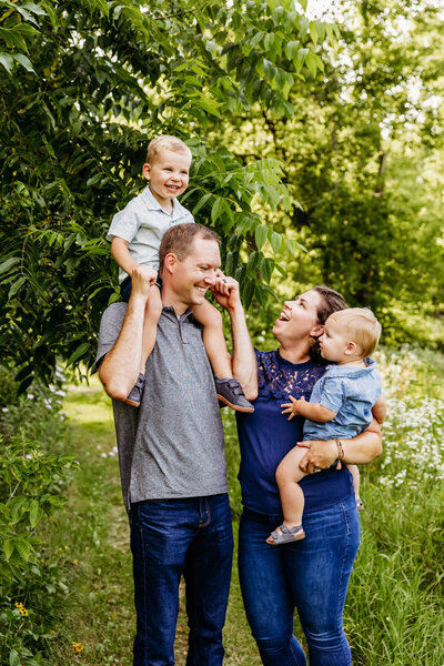 dad walking with his toddler son on his shoulders as they look at mom who's carrying their youngest son