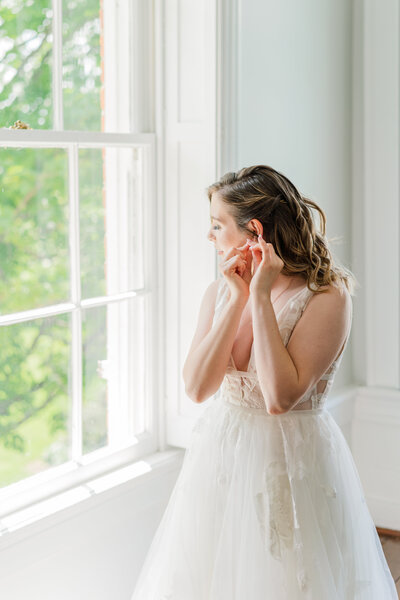 Older bride in fitted lace dress with long sleeves smiles and looks out the window as her daughter buttons up the back of her wedding dress