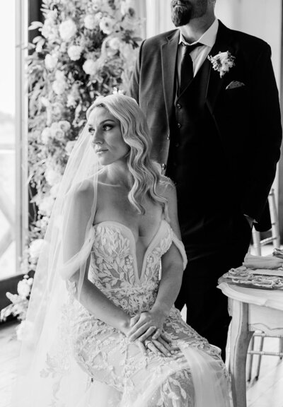 Bride sitting with groom standing behind her in black and white