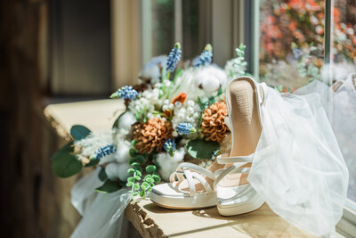 Photo of bride’s elegant white wedding shoes, her delicate veil draped over a vibrant bouquet of rust floral and greenery.