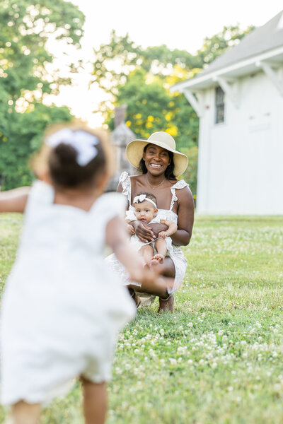 A photoshoot of a mother and two daughters playing in the field. The mother is bent down while holding the baby as her older daughter runs towards them.