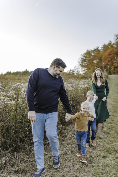 Verona B Photography captures a beautiful fall family photo of a mom, dad, and their two boys at Kendall Hills in Peninsula, OH. The family are holding hands and walking along a hiking path down a hill with trees in the background. The fall colors were really at peak color and the sun was perfect for this family photo session near Akron.