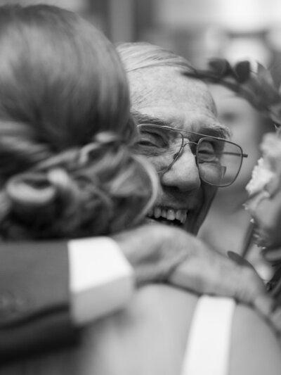 A grandpa smiles over his granddaughter's shoulder as she hugs him at her wedding.