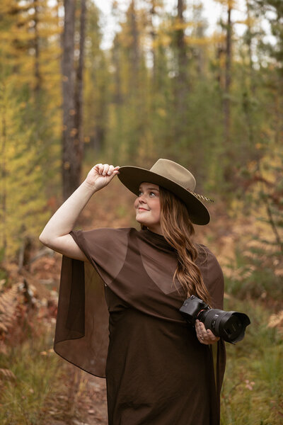 Sydney Breann, a Montana-based elopement photographer, smiles while holding her camera and adjusting her hat on a wooded trail surrounded by autumn colors.