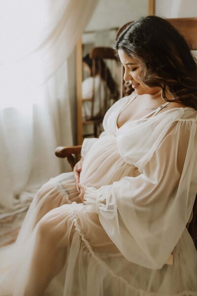 Pregnant woman in a white sheer gown sits on a wooden chair, cradling her belly and looking down, with soft natural light filtering through a nearby window.