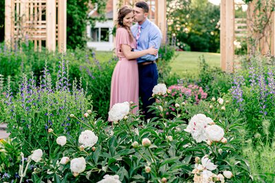 An engaged couple shares an embrace in the background as the camera focuses on beautiful white peonies in bloom during a spring engagement session at Green Spring Gardens Park in Northern Virginia