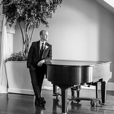 B&W Utah wedding photography of a groom leaning against a black grand piano in the clubhouse at Sleepy Ridge Golf Course, Orem