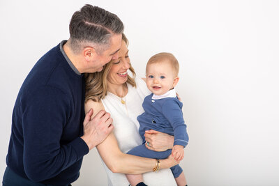 sweet family of three with dad hugging mom while she holds their toddler who is looking at the camera