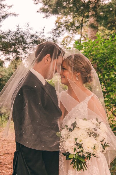 Bride and groom share an intimate moment under a veil in a forest, holding a bouquet of white flowers, celebrating their union with one of our stress-free wedding packages.