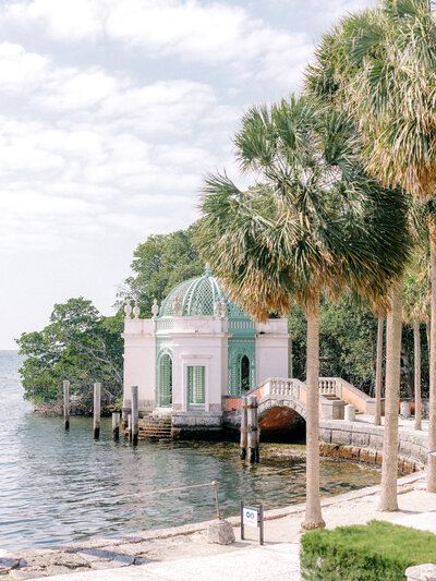 Pink and green boathouse by the water with palm trees in the foreground