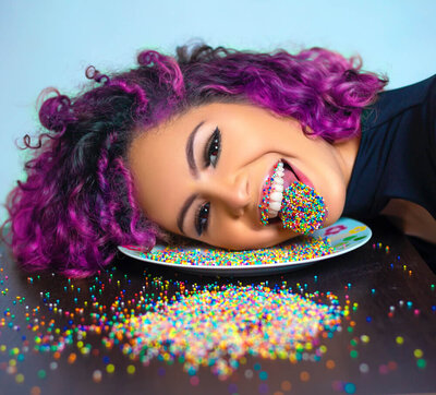 Creative woman with purple hair laying her head in a plate of sprinkles, with sprinkles on her tongue