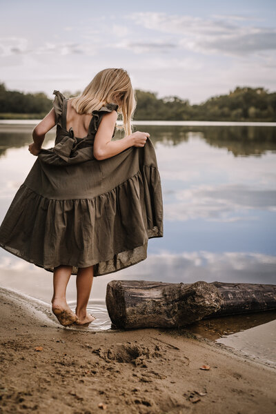 Young girl in an olive-green dress standing by a lake, holding her dress.