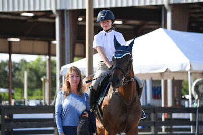 girl posing on her horse in horse show attire with her mother smiling next to her in a blue shirt