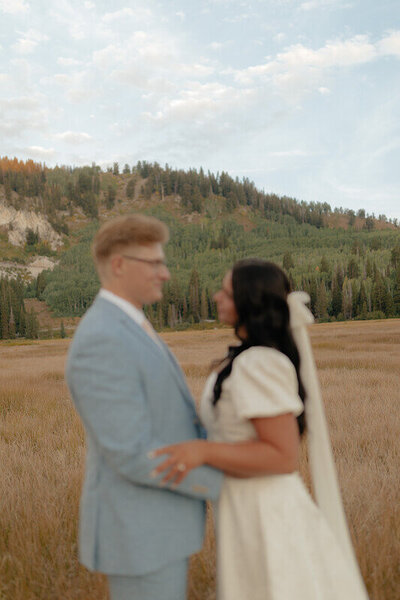 A bride and groom hold each other by the waist. The are blurred with the hill with trees behind them in focus.
