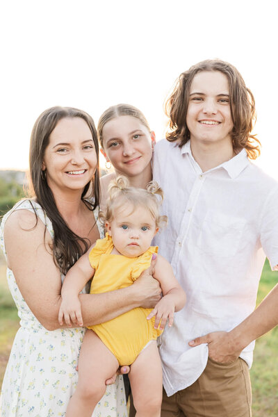 Mum holds toddler girl dressed in daffodil yellow while teenage brother and sister stand cuddled in close by. Photo taken at Cooee Bay, Yeppoon.