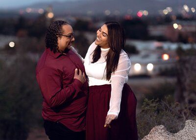 Man in red business shirt and woman in red skirt showing off her ring with city lights in background.