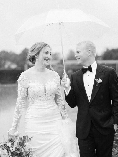 Black and white photo of bride and groom smiling at each other under an umbrella